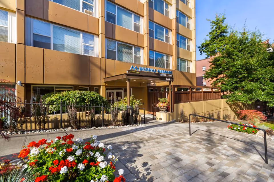 Exterior view of a multi-story senior living facility with a brown and beige facade, large windows, and an entrance canopy. The entrance is surrounded by greenery, including bushes and colorful flowers, with a paved walkway leading up to the door. A tree and additional plants are visible on the right side under a clear blue sky.