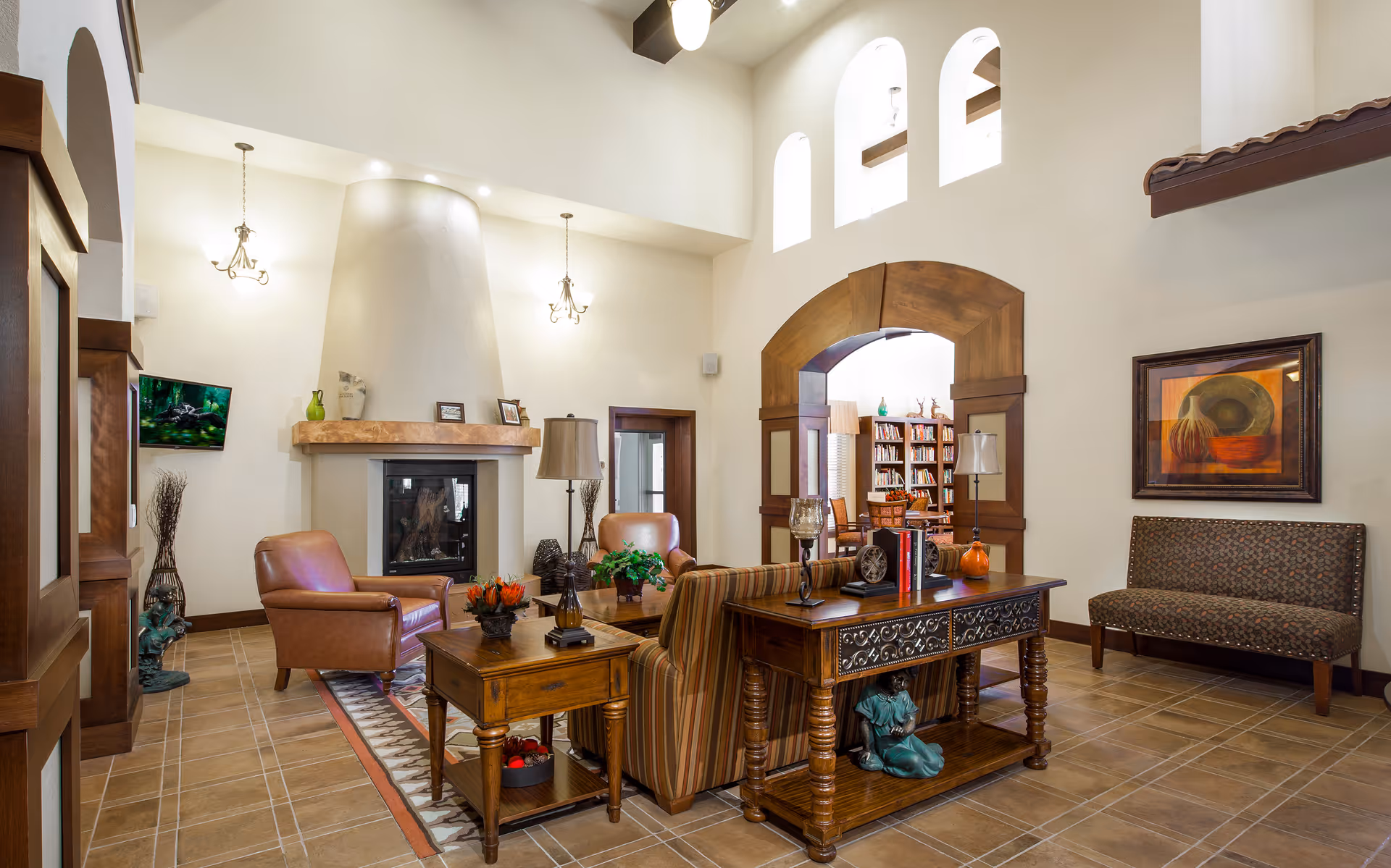 A cozy senior living common area with a fireplace, leather armchairs, a striped sofa, wooden tables, and decorative items. The room features high ceilings with arched windows and an open doorway leading to a room with bookshelves and a table with chairs.