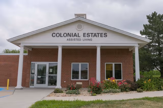 Front exterior view of a brick building with white columns and a triangular pediment. The building has a sign that reads 'COLONIAL ESTATES ASSISTED LIVING'. There are windows, a glass door entrance, and landscaping with flowers and shrubs in front.