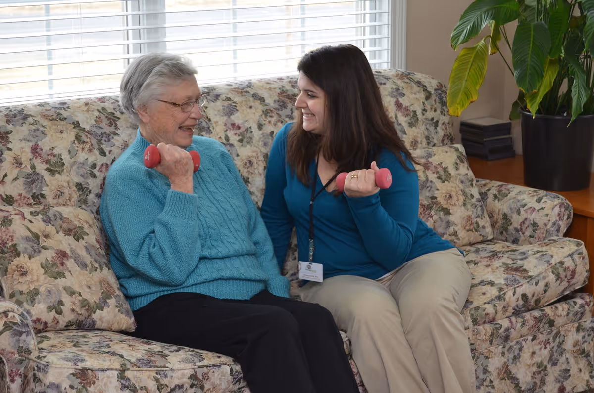 An elderly woman and a younger woman sitting on a floral-patterned couch, both holding small pink dumbbells and smiling at each other in a bright room with a large window and a potted plant nearby.