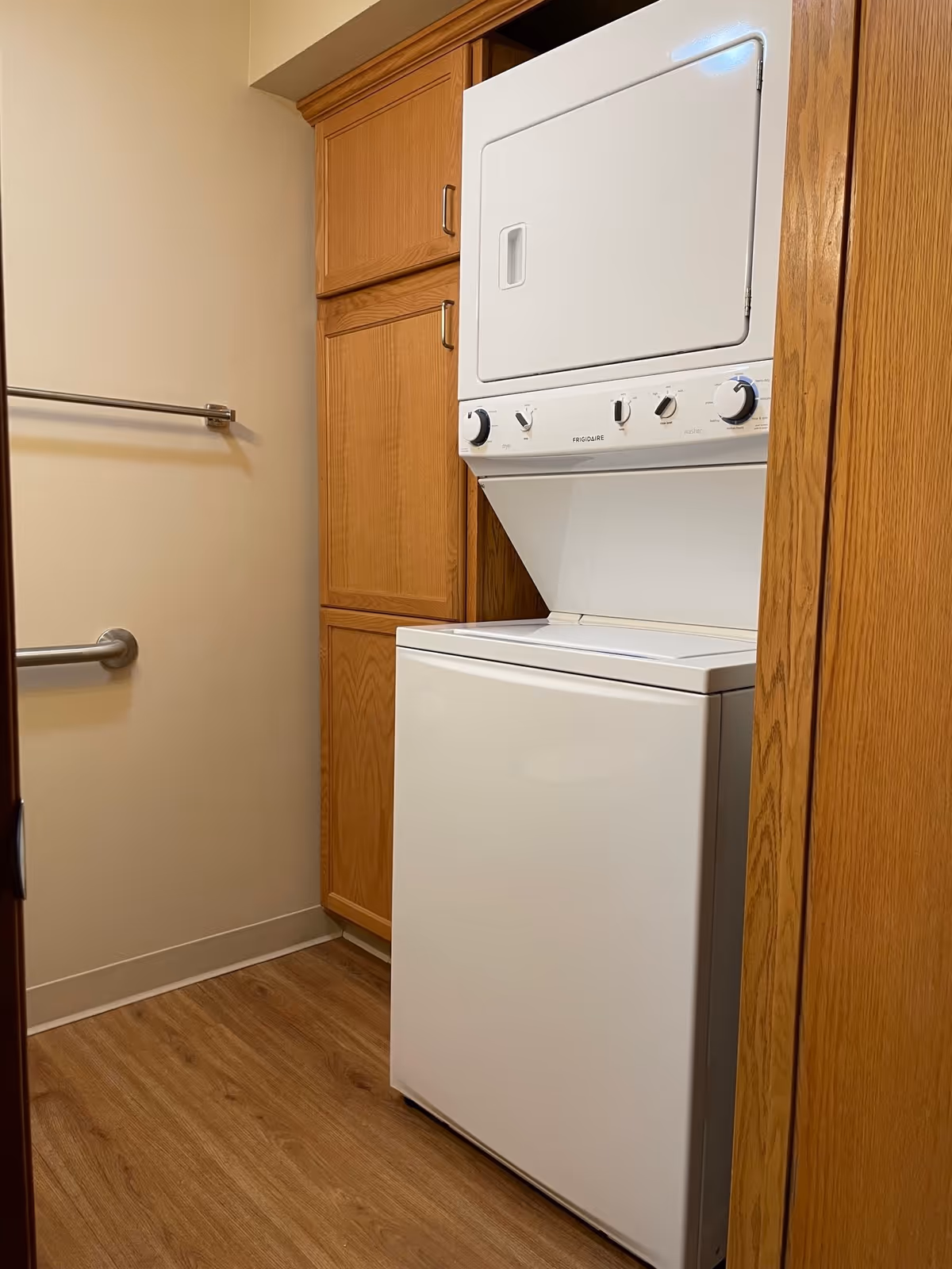 A small laundry area with a stacked white washer and dryer unit next to wooden cabinets. The floor is wood-patterned, and there are two metal grab bars mounted on the beige wall.
