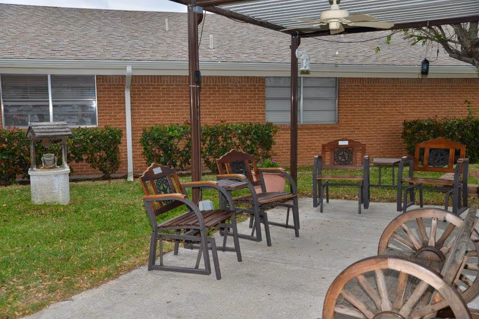 Outdoor seating area with wooden and metal chairs arranged under a pergola with a ceiling fan. The area is adjacent to a brick building with windows and surrounded by green grass and bushes. A decorative wishing well is visible on the left side of the image.