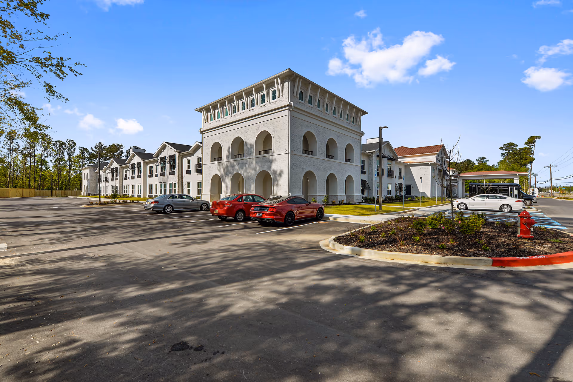 Exterior view of The Blake at Biloxi senior living facility showing a large white building with arched balconies, several parked cars in the parking lot, landscaped areas with small plants, and a clear blue sky with some clouds.