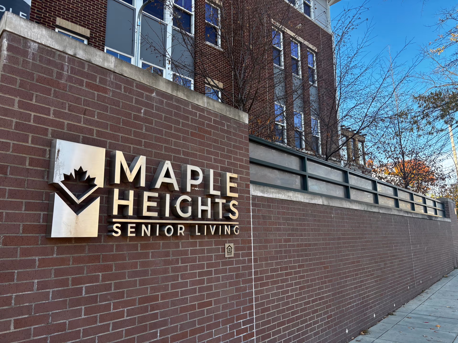 Exterior view of a brick wall with a metal sign that reads 'Maple Heights Senior Living' attached to it. Behind the wall, there is a multi-story brick building with several windows and some leafless trees. The sky is clear and blue.