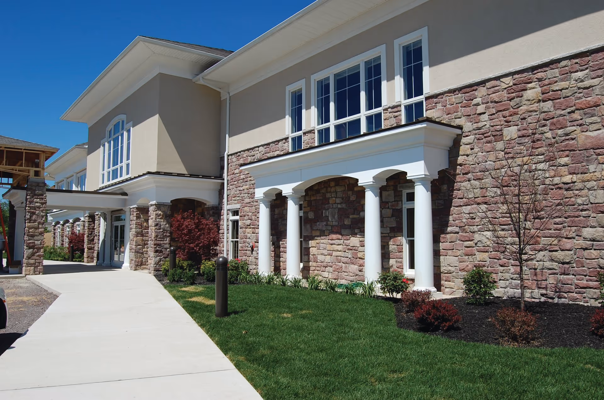 Entrance and front exterior of a stone-faced senior living building with white columns, windows, and a sidewalk.