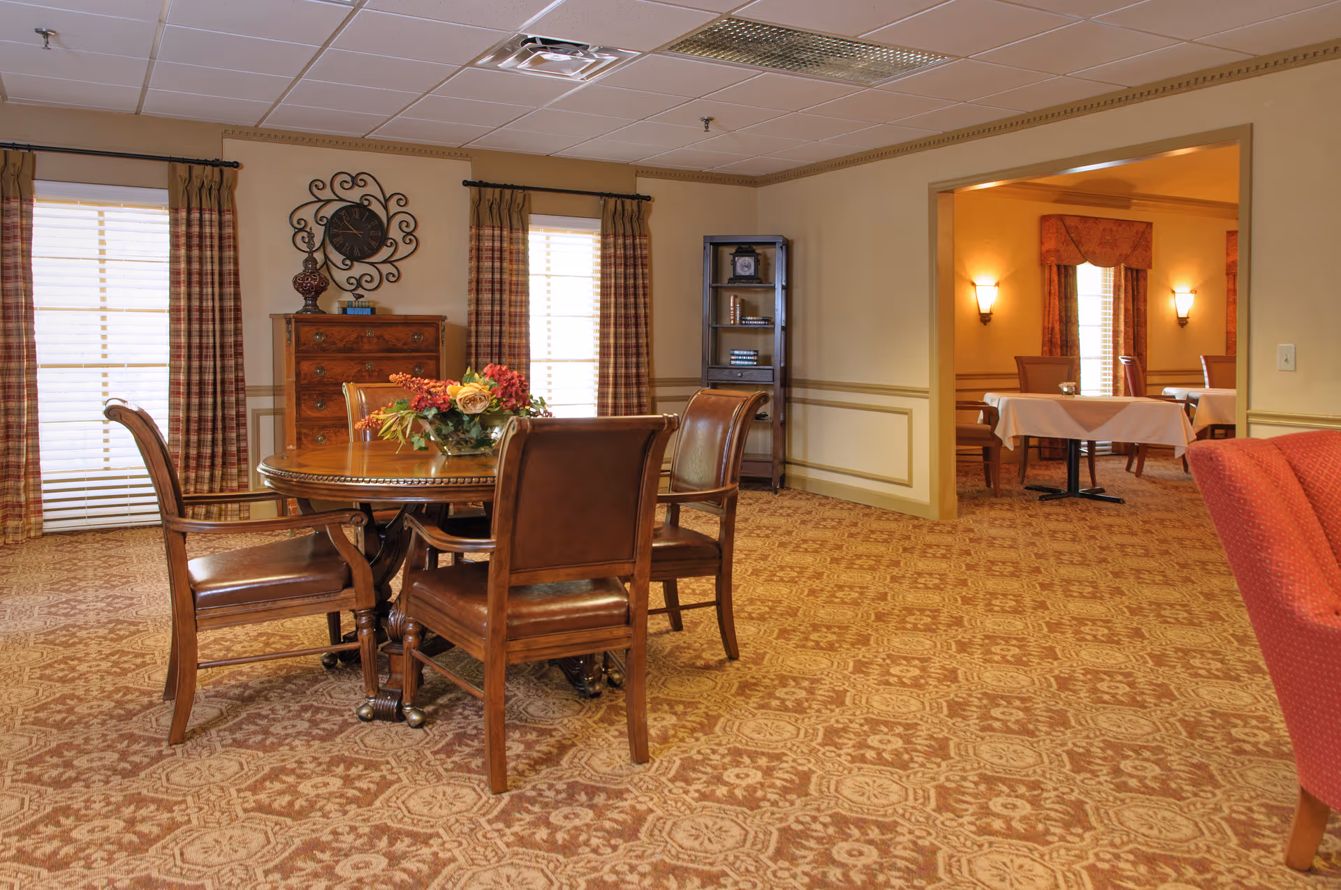 Warmly decorated senior living dining/lounge area with a round wooden table and chairs, patterned carpet, and a doorway to another dining room.