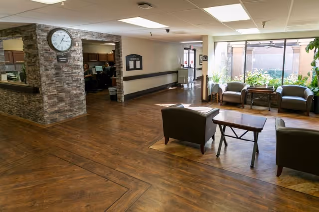 Spacious reception lobby with wood floors, chairs and small tables, a stone-clad reception desk with a clock, and a large window overlooking a garden.