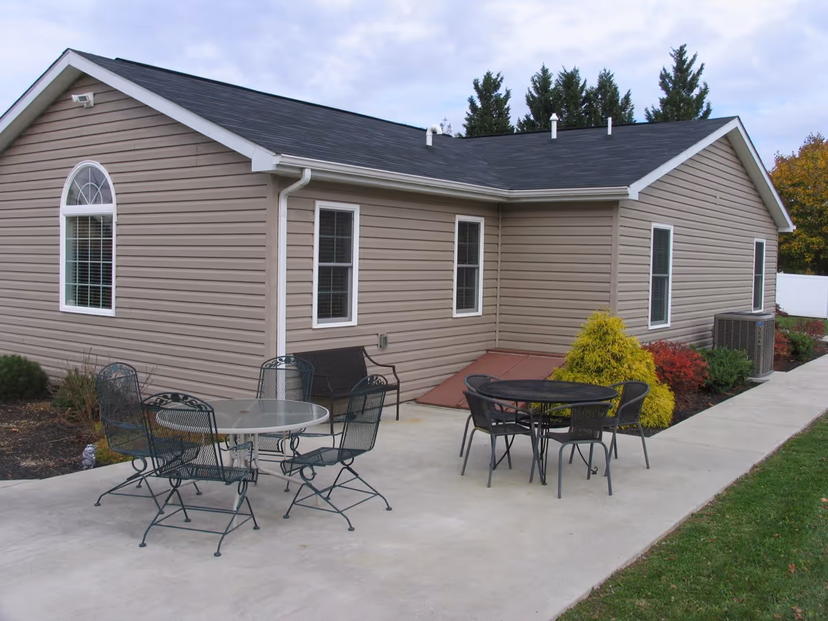 Exterior view of a beige single-story building with metal patio tables and chairs on a concrete walkway and surrounding shrubs.