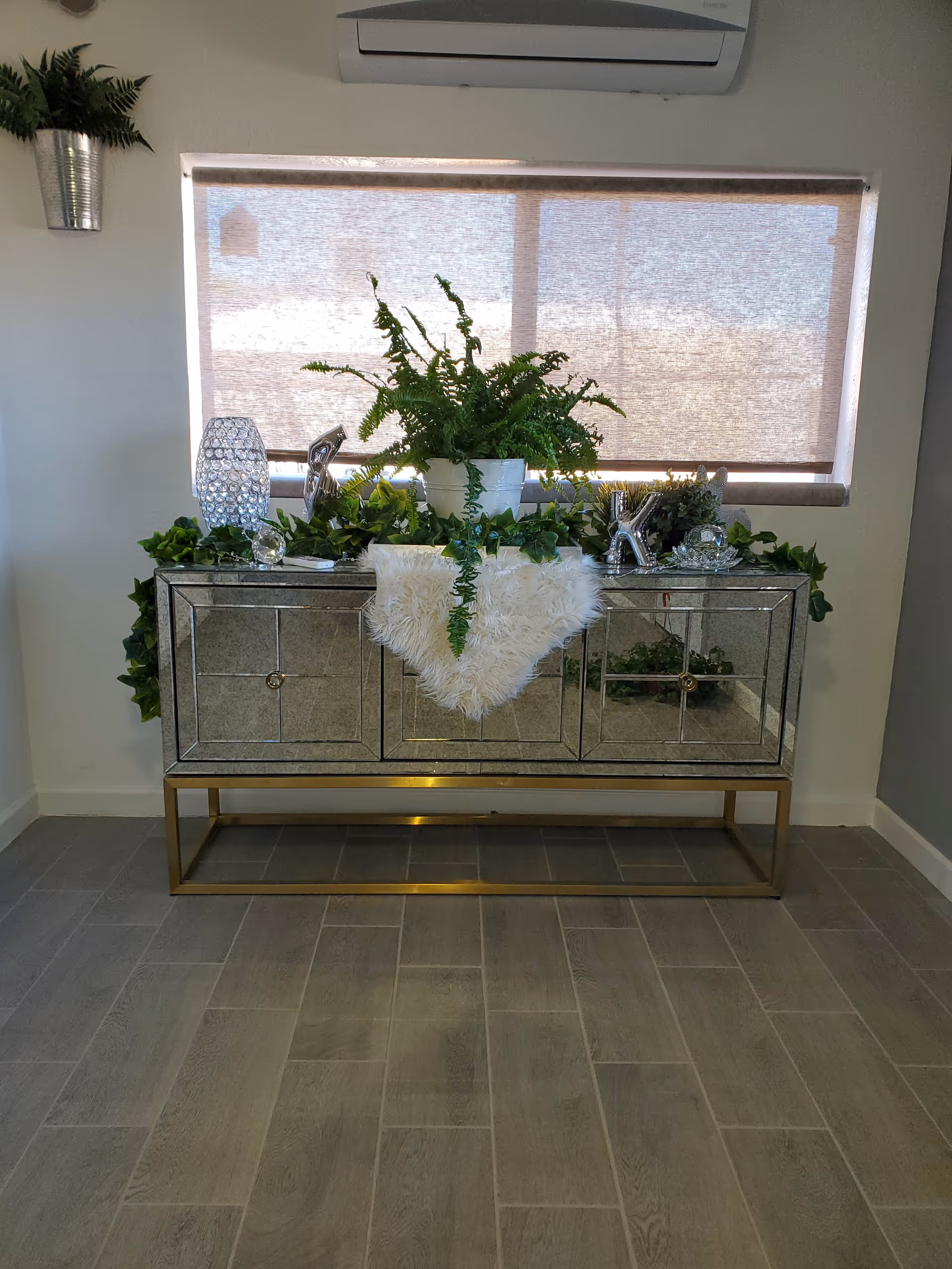 A mirrored console table with a gold metal frame stands against a wall beneath a window with a beige roller shade. The table is decorated with green plants, a white fluffy cloth, and crystal-like decorative items. A small potted plant is mounted on the wall to the left, and an air conditioning unit is installed above the window. The floor is covered with gray wood-look tiles.