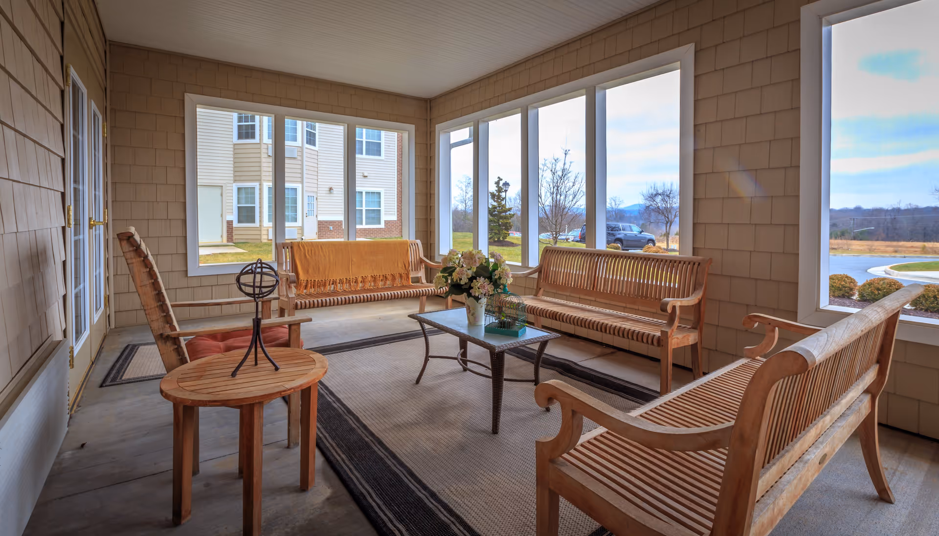 A bright enclosed porch area with wooden benches and chairs arranged around a rectangular glass-top coffee table. The porch has large windows offering views of the outdoors, including a lawn, trees, and a building in the background. A decorative centerpiece with flowers is on the coffee table, and a small round wooden side table with a decorative object is also present.