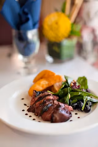 A plated meal featuring sliced grilled steak drizzled with sauce, accompanied by a small salad with leafy greens and black olives, and a serving of roasted potato wedges. The background shows a blurred table setting with a blue napkin and a yellow flower in a green vase.
