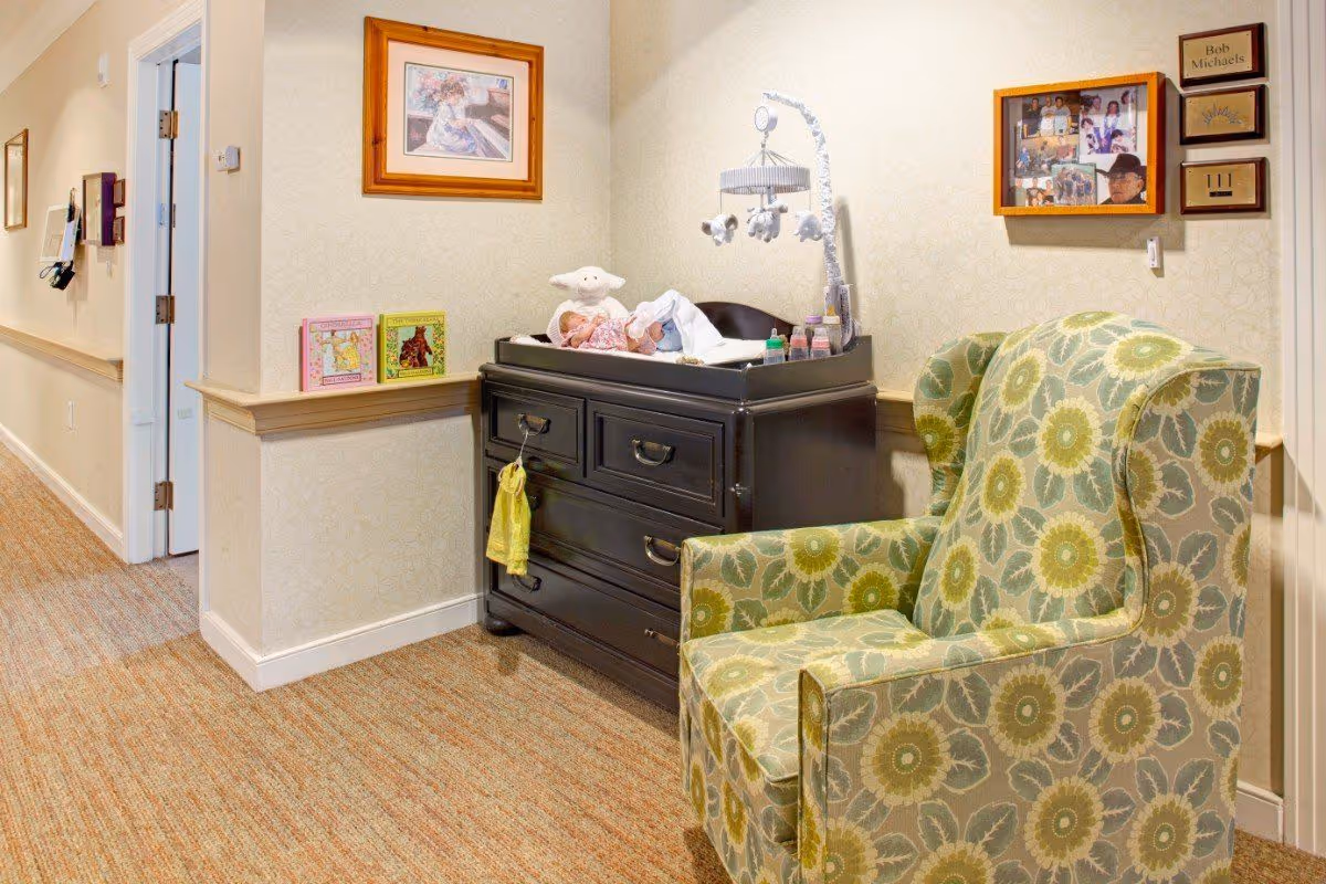 A hallway seating area with a green floral armchair beside a dark dresser topped with baby items and framed pictures on the wall.