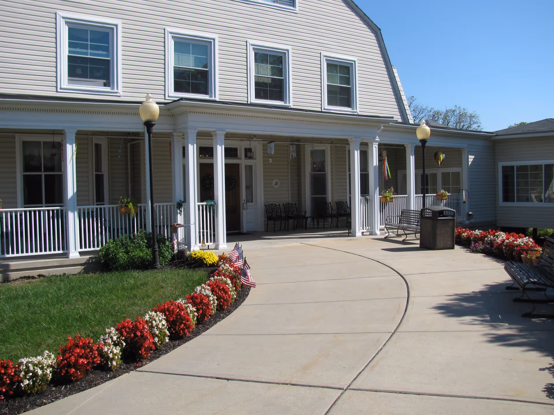 Front entrance of Ridgeway Village Assisted Living with a curved concrete walkway, flower beds with red and white flowers, American flags, benches, lamp posts, and a porch with white columns and chairs.