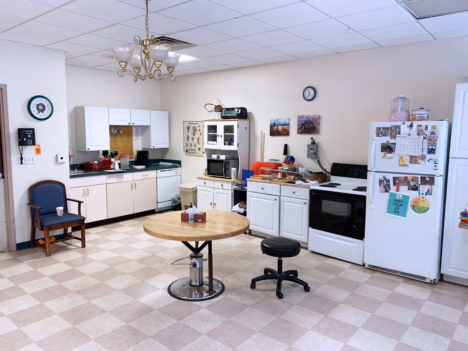 A kitchen area in a rehabilitation and nursing facility featuring white cabinets, a white refrigerator covered with various magnets and photos, a white stove, a microwave on a cabinet, a round wooden table with a tissue box on it, a black rolling stool, and a blue upholstered chair. The room has a checkered tile floor, a chandelier light fixture, and a wall clock.