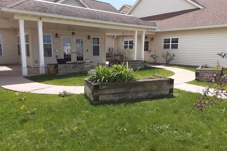 Outdoor courtyard area at Milestone Senior Living Eau Claire featuring a green lawn with raised wooden garden beds containing plants, a curved concrete walkway, and a beige building with multiple windows and doors under a covered porch.
