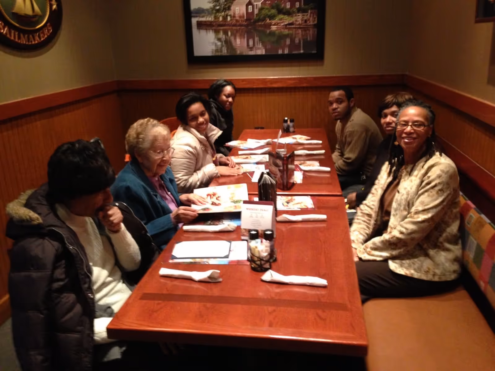 A group of seven people sitting around a wooden dining table in a cozy restaurant booth, looking at menus and smiling. The walls have wood paneling and framed pictures.
