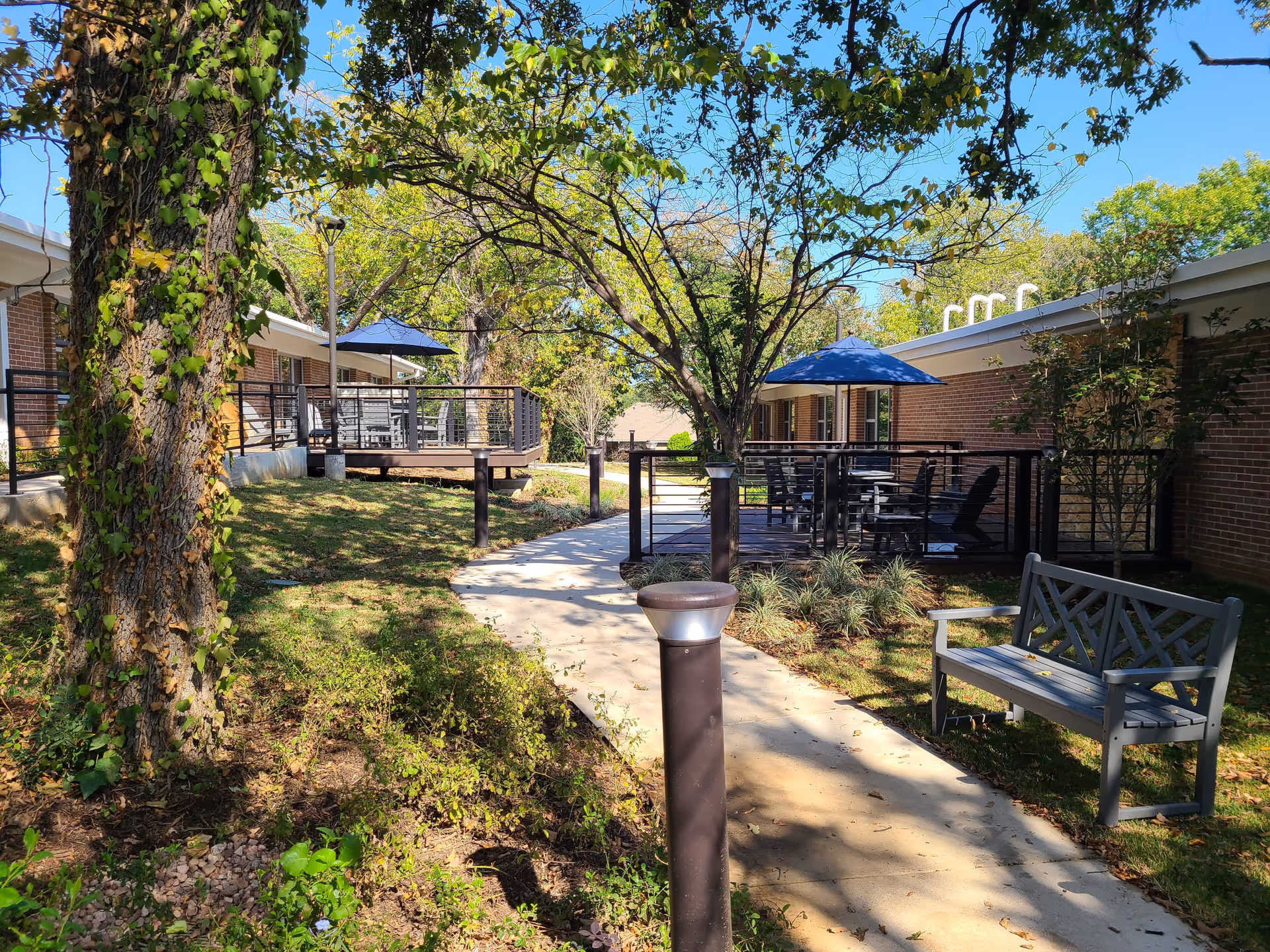 Outdoor patio area at Fairhaven Denton with a paved walkway, trees, and greenery. There are two raised decks with tables, chairs, and blue umbrellas. A gray bench is positioned near the walkway under the shade of a tree. The building exterior is brick with white trim.
