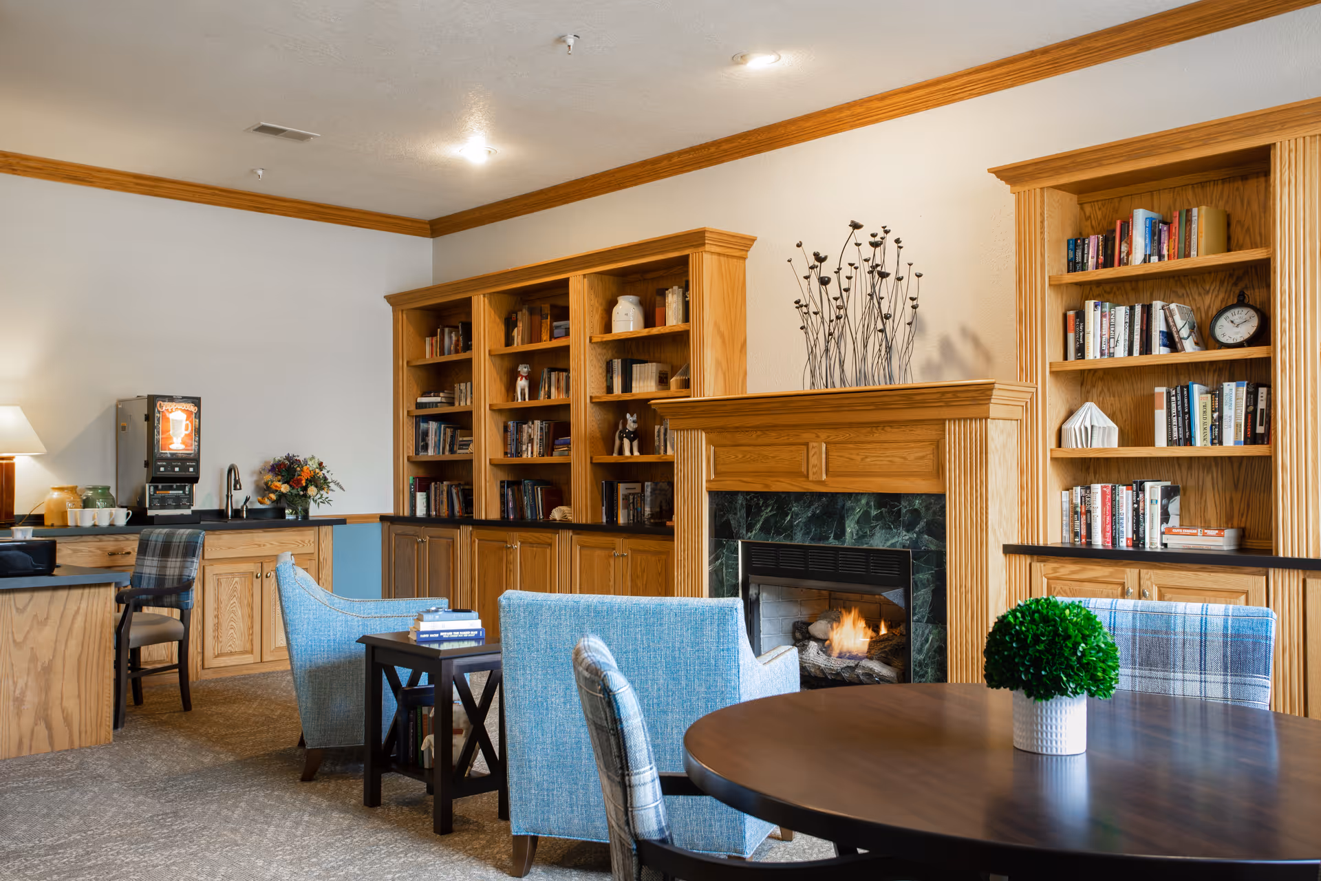 A cozy communal living room with bookshelves, a fireplace, upholstered chairs and a round table in a senior residence.