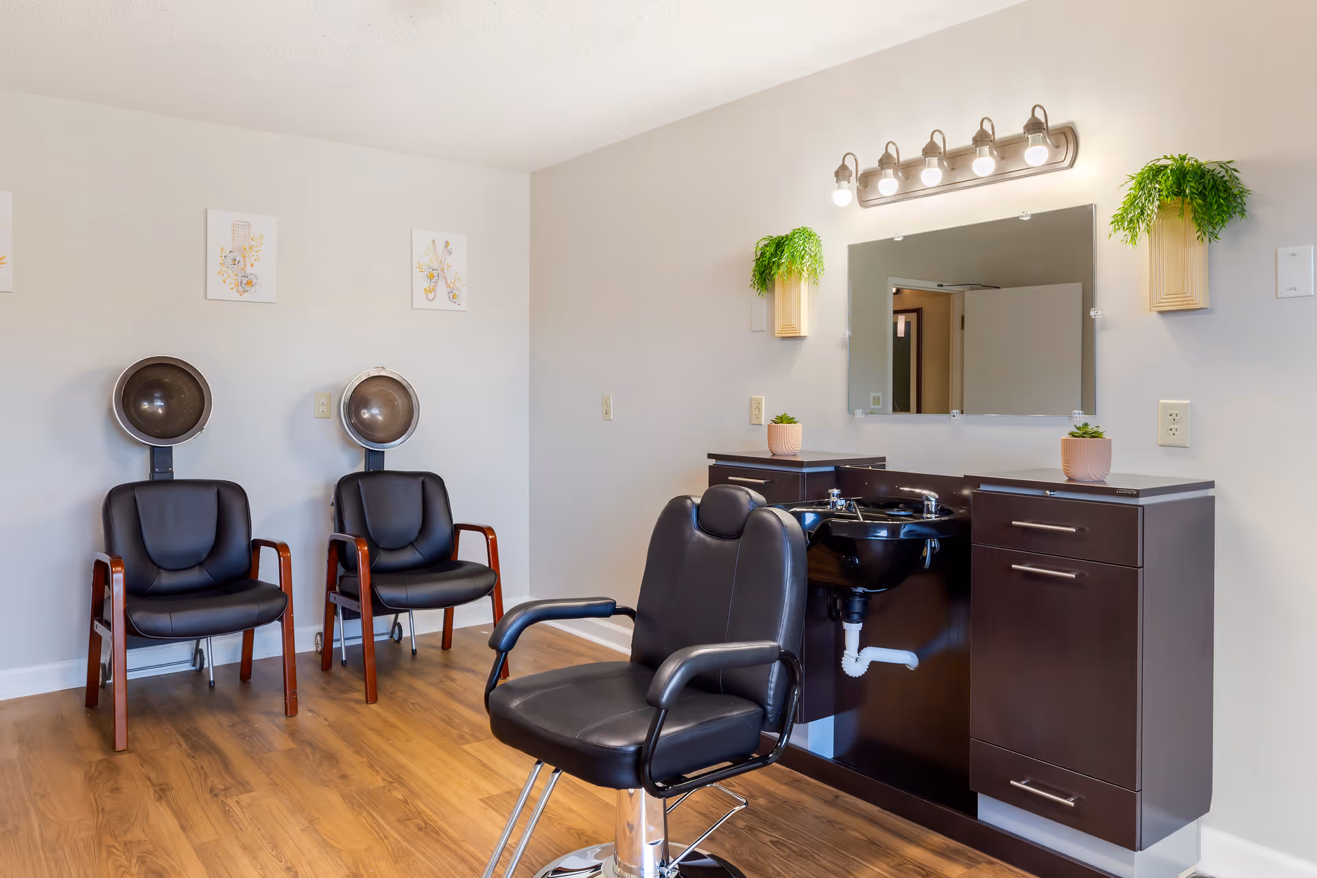 Interior of a salon area in a senior living facility with two black leather chairs under hair dryers against a wall, a black salon chair in front of a black sink and cabinet, a large mirror with lights above it, and two small potted plants on the cabinet and wall-mounted planters.