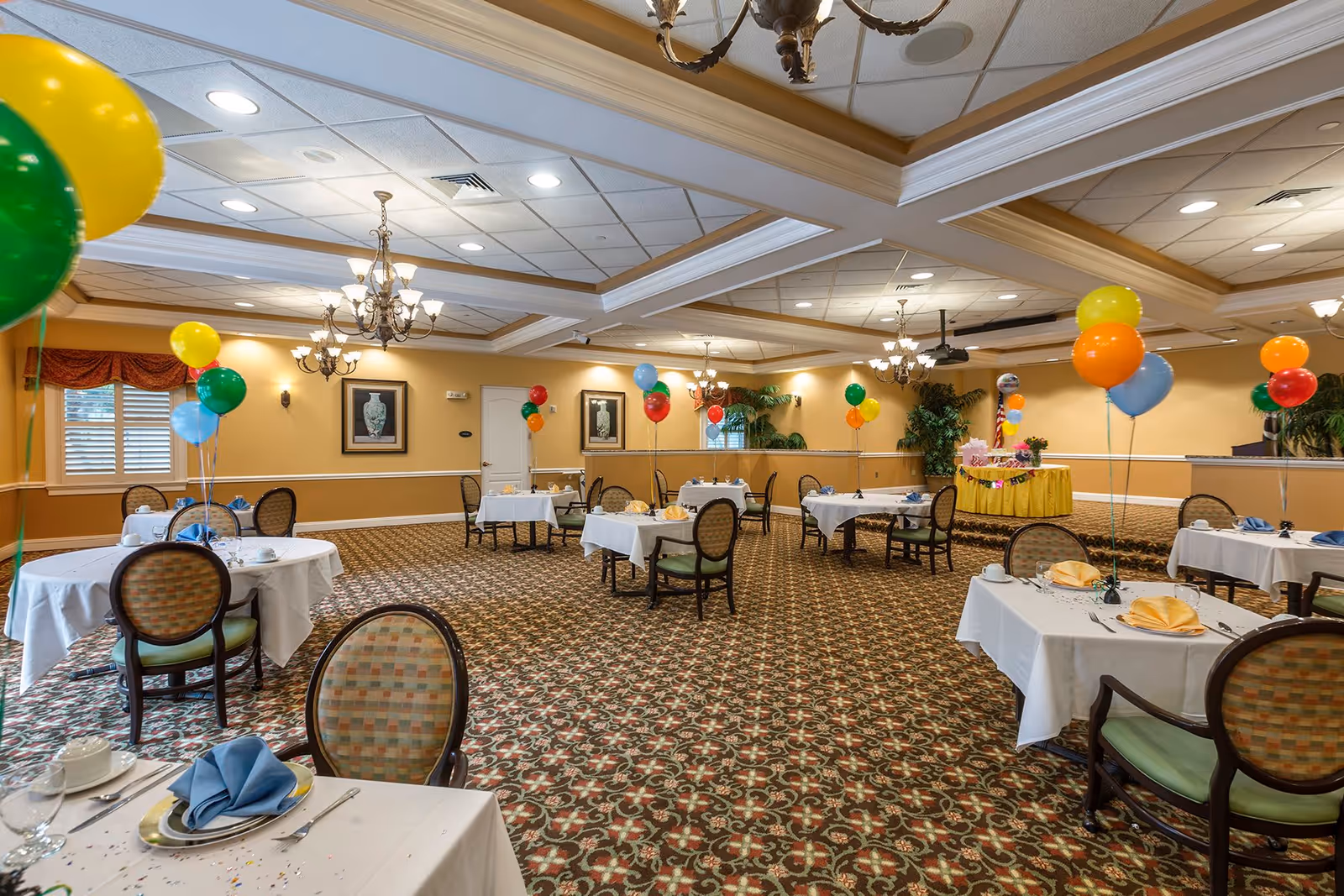 A spacious dining room decorated with colorful balloons tied to chairs and tables set with white tablecloths, plates, napkins, and silverware. The room features patterned carpet, chandeliers, framed artwork on the walls, and a small stage area with a yellow tablecloth and more balloons.