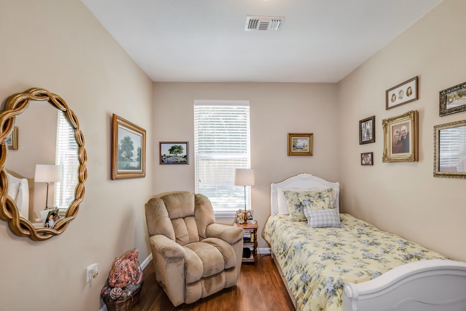 Small furnished bedroom with a single floral bed, recliner chair, bedside table and framed pictures on the walls.