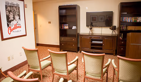 A small media room with six beige and wood-framed chairs arranged in two rows facing a wall-mounted flat screen TV. Below the TV is a wooden entertainment console with decorative items and two tall wooden bookshelves on either side, one holding DVDs and books. A framed Casablanca movie poster is hung on the left wall. The room has beige walls and a patterned carpet.