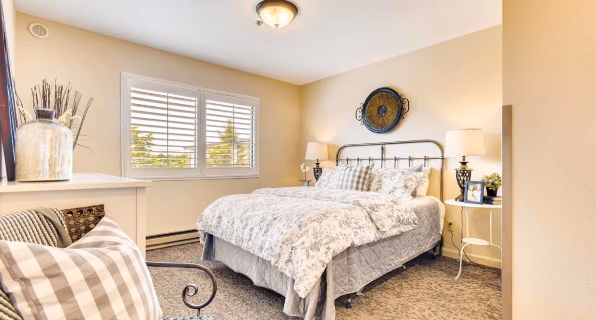 A cozy bedroom with a large bed covered in floral patterned bedding and multiple pillows. There are two bedside tables with lamps on each side of the bed. A decorative wall piece hangs above the bed. A window with white shutters lets in natural light. A chair with a plaid cushion is partially visible in the foreground, and a dresser with decorative items is on the left side of the room.