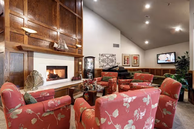 A cozy living room area with floral patterned red armchairs arranged around a glass coffee table in front of a lit fireplace with wooden paneling above it. The room has a high ceiling with recessed lighting, a wall-mounted TV, a piano, decorative wall art, and a large potted plant in the corner.