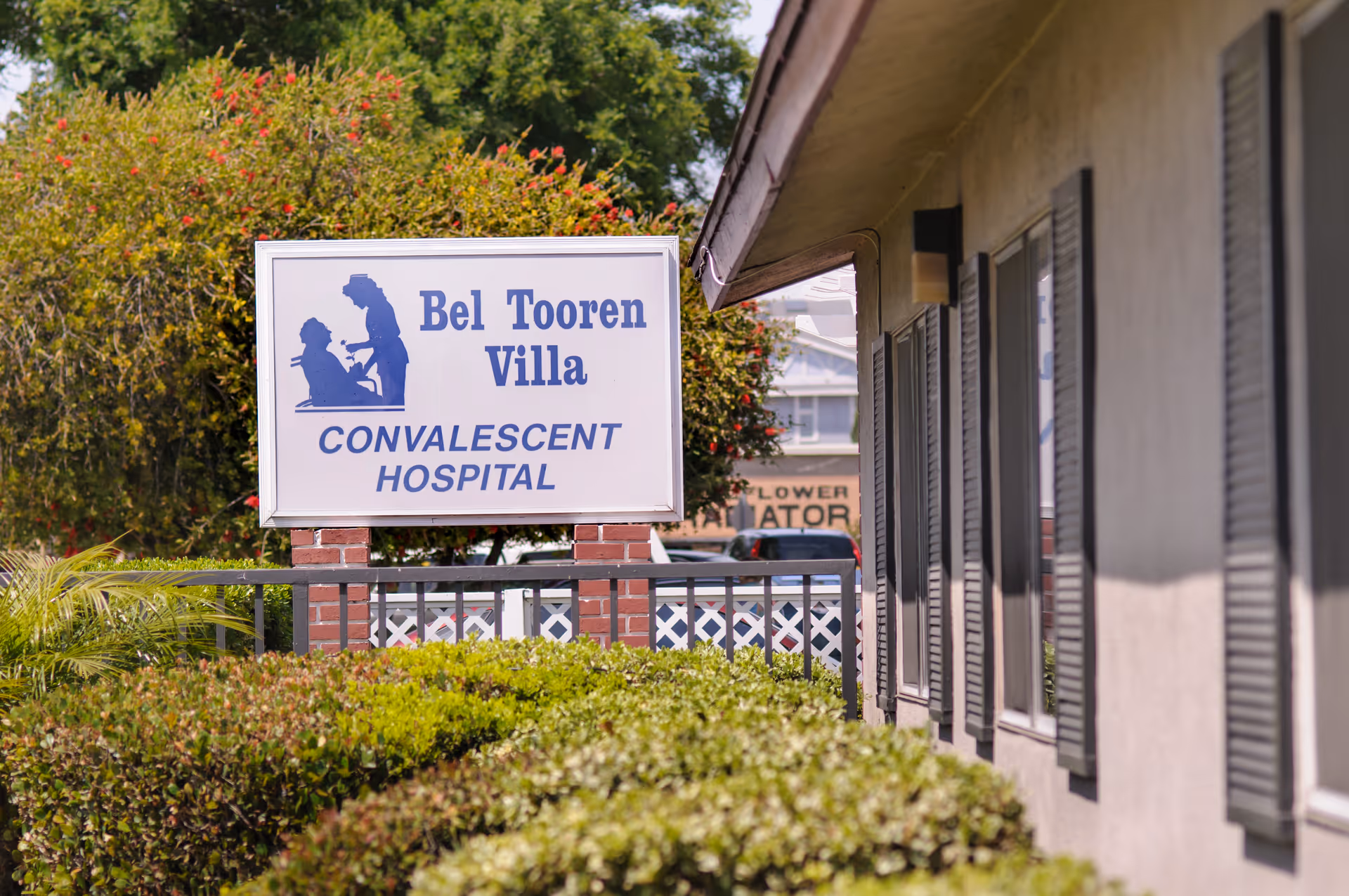 Outdoor view of the Bel Tooren Villa Convalescent Hospital sign with bushes and greenery in the foreground and part of the building with windows and shutters on the right side.