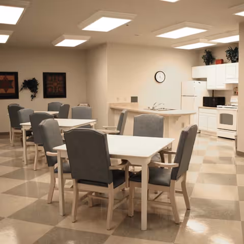 Interior view of a communal dining area with several tables and cushioned chairs. The room has a checkered floor, white walls, and a kitchen area with white cabinets, a refrigerator, microwave, and stove. There are framed artworks and a plant on the wall, and a clock above the kitchen counter.