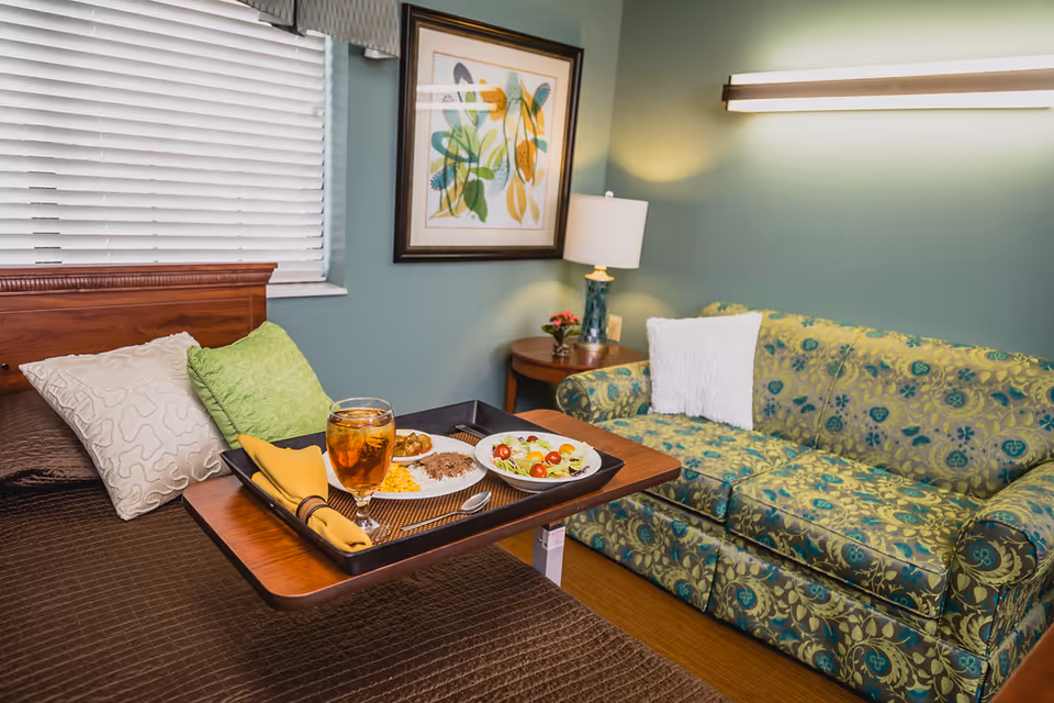 A cozy room with a brown bed featuring two pillows, a green and a white one. A wooden tray table on the bed holds a meal with a glass of iced tea, a plate with corn, meat, and vegetables, and a bowl of salad. Next to the bed is a green patterned couch with a white pillow, a wooden side table with a lamp and a small plant, and a framed botanical artwork on the wall. The room has green walls and a window with white blinds.