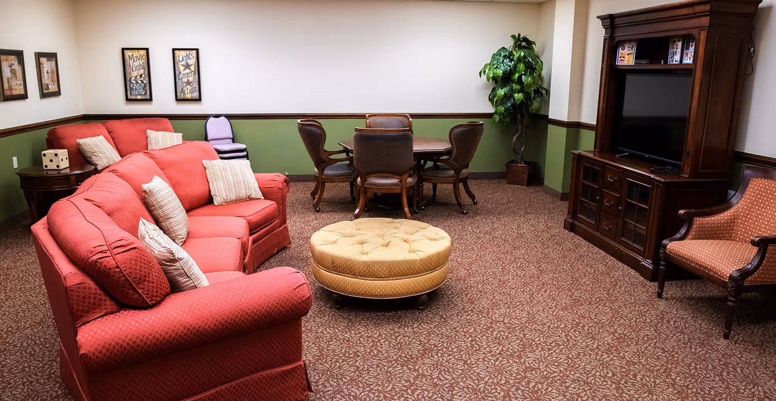 A cozy living room with red upholstered sofas and striped pillows, a round beige ottoman in the center, a wooden entertainment center with a TV, a wooden table with four leather chairs, a potted plant in the corner, and framed wall art with movie-themed quotes.