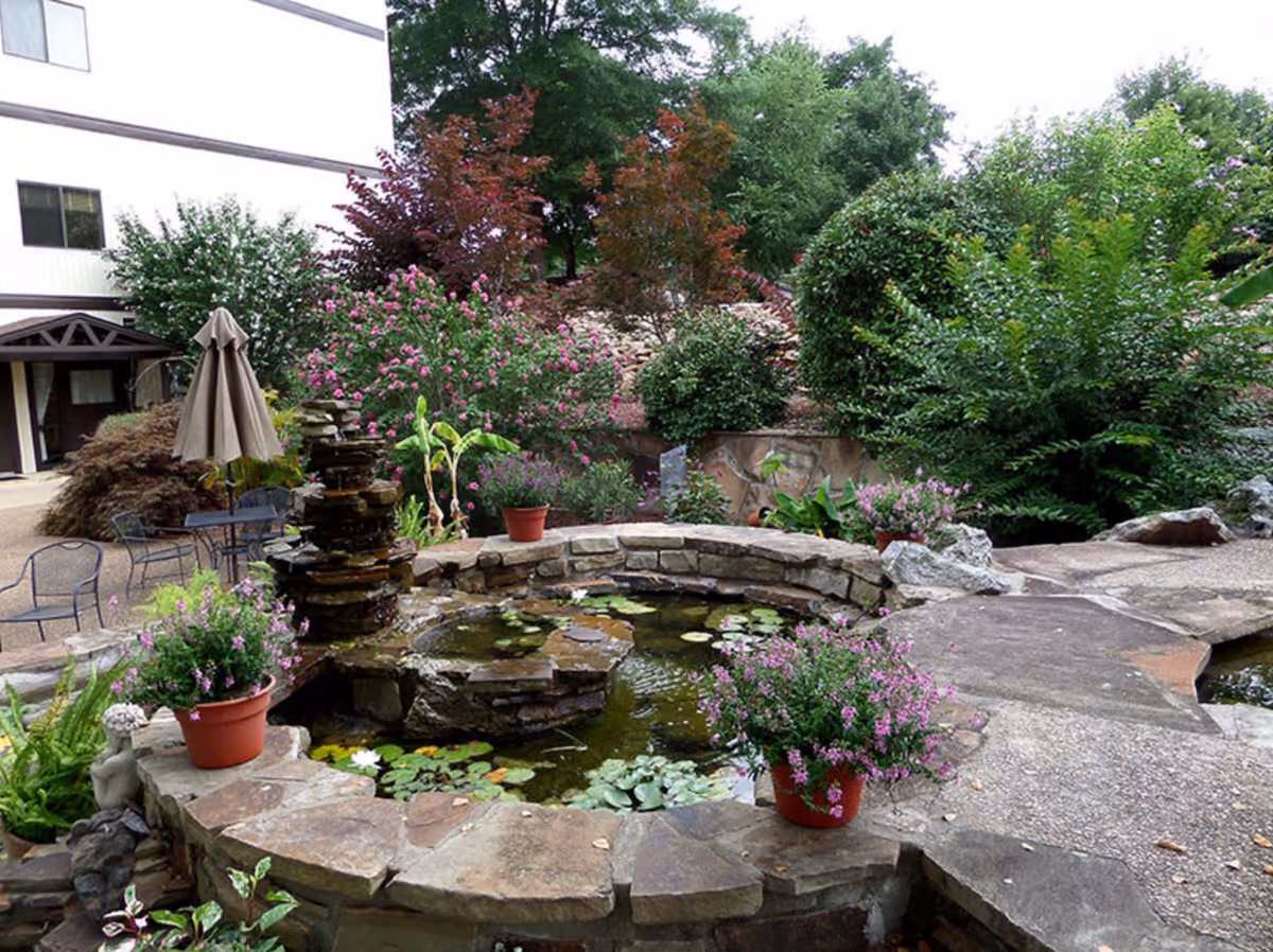 Landscaped outdoor courtyard with a stone pond and fountain surrounded by potted flowers, patio seating, and lush greenery beside a building.