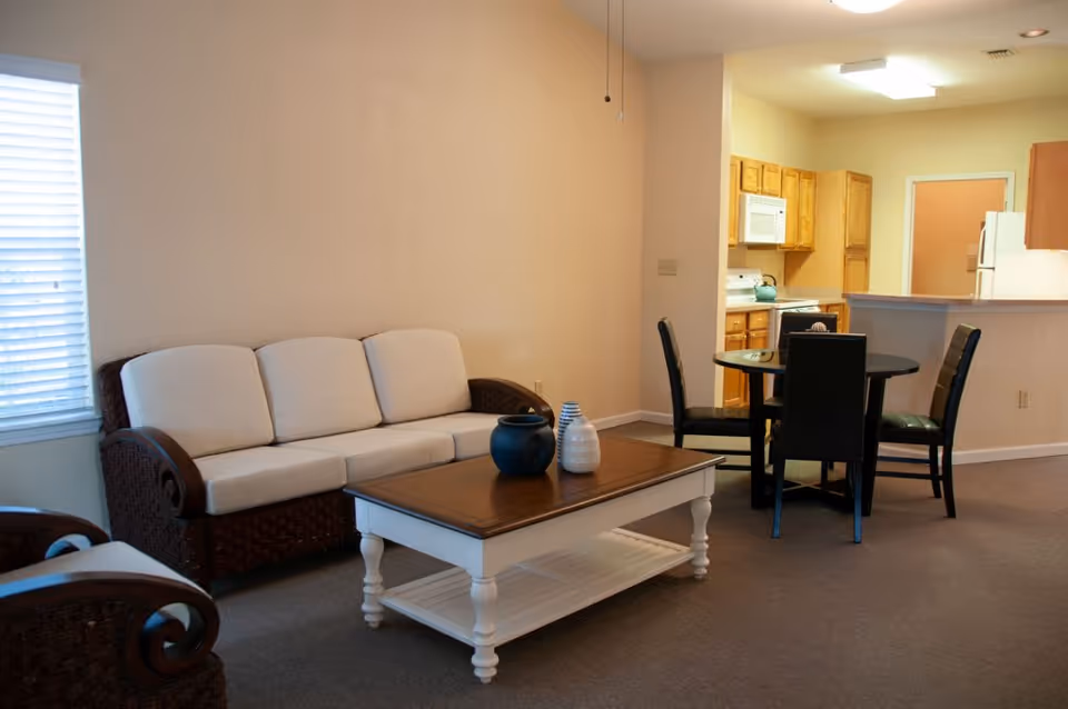 Open living room with a white-upholstered wicker sofa, a coffee table with decorative vases, a small dining table with chairs, and an adjacent kitchen.