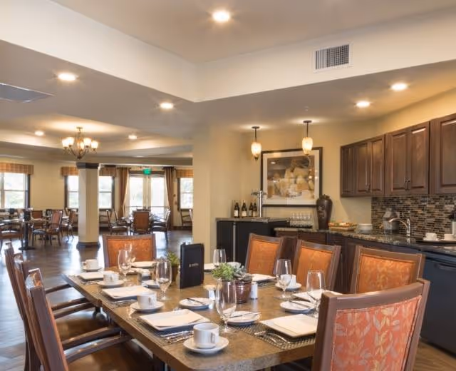 A spacious dining room in a senior living facility with a large table set for a meal, including plates, cups, glasses, and napkins. The room features wooden chairs with orange patterned cushions, a kitchenette area with dark wood cabinets, a tiled backsplash, and a countertop with wine bottles and glasses. Large windows with curtains allow natural light to fill the room.