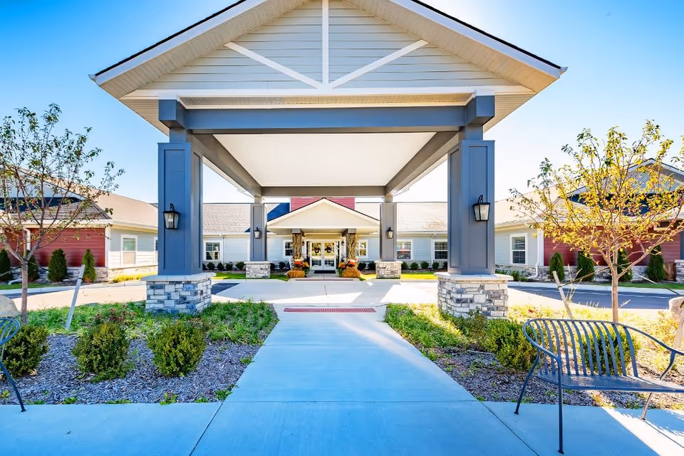 Front entrance of Provision Living at Fenton, featuring a covered driveway supported by large pillars with stone bases, landscaped greenery, and benches on either side of the walkway leading to the glass doors of the building.