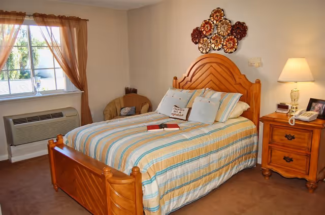 A cozy bedroom with a wooden bed frame and a striped bedspread in shades of blue, beige, and white. There are several pillows on the bed, including one with the text 'Carrots are a vegetable.' A small book and glasses rest on the bed. To the right of the bed is a wooden nightstand with a lamp, a telephone, and a framed photo. A decorative metal wall art piece hangs above the headboard. A window with sheer brown curtains lets in natural light, and a beige armchair is placed in the corner near the window. The room has beige walls and carpeted flooring.