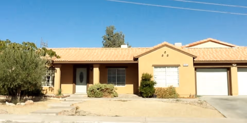 Single-story stucco house with a tiled roof, front door, garage, and a dry landscaped yard under a clear blue sky.