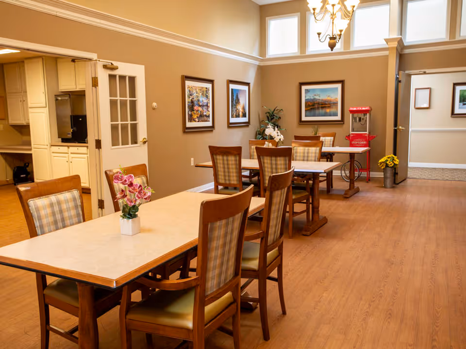 A dining area in a senior living facility with wooden tables and chairs featuring plaid upholstery. The room has beige walls adorned with framed landscape paintings, a popcorn machine in the corner, and a vase with flowers on one of the tables. The floor is wood, and there is a doorway leading to a kitchen area with white cabinets.