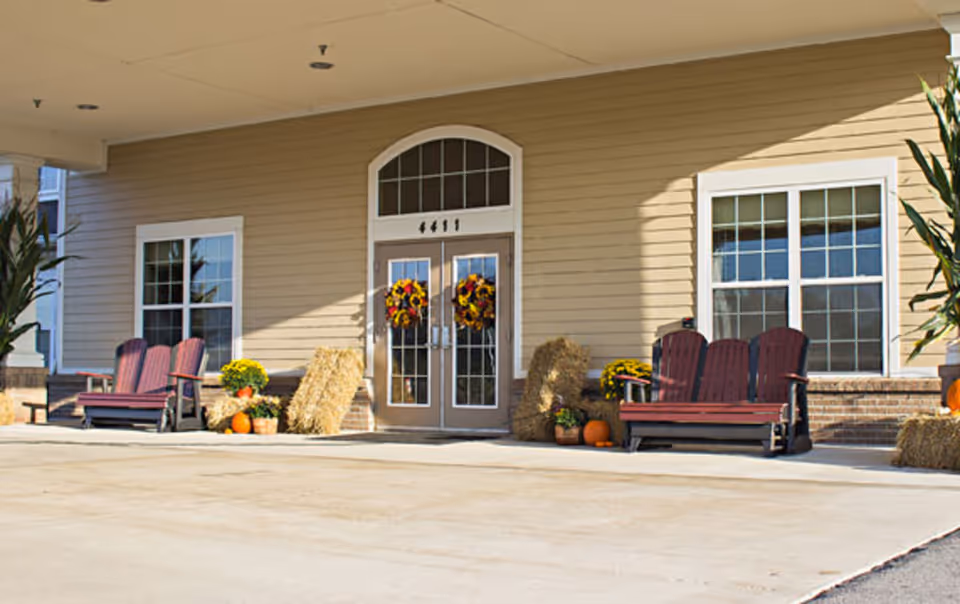 Entrance of a senior living facility with double glass doors decorated with autumn wreaths. There are two sets of red wooden benches on either side of the entrance, surrounded by hay bales, pumpkins, and potted plants. The building exterior is beige with white trim around the windows and door.
