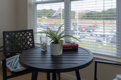 A small round black table with a potted fern plant, a glass, and two stacked books on it, positioned in front of a window with white blinds. Two black chairs with patterned cushions are partially visible around the table. Outside the window, a parking lot and a flagpole with a flag can be seen.