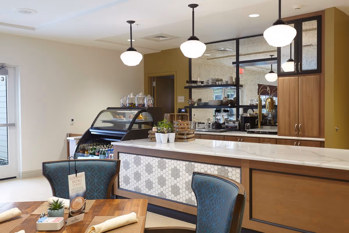 A modern dining area with a wooden table set with napkins and a small plant. Behind the table is a counter with a white marble top and hexagonal tile design on the front. Above the counter hang three pendant lights. Behind the counter are shelves with dishes and a refrigerated display case with beverages. The room has a warm and inviting atmosphere.