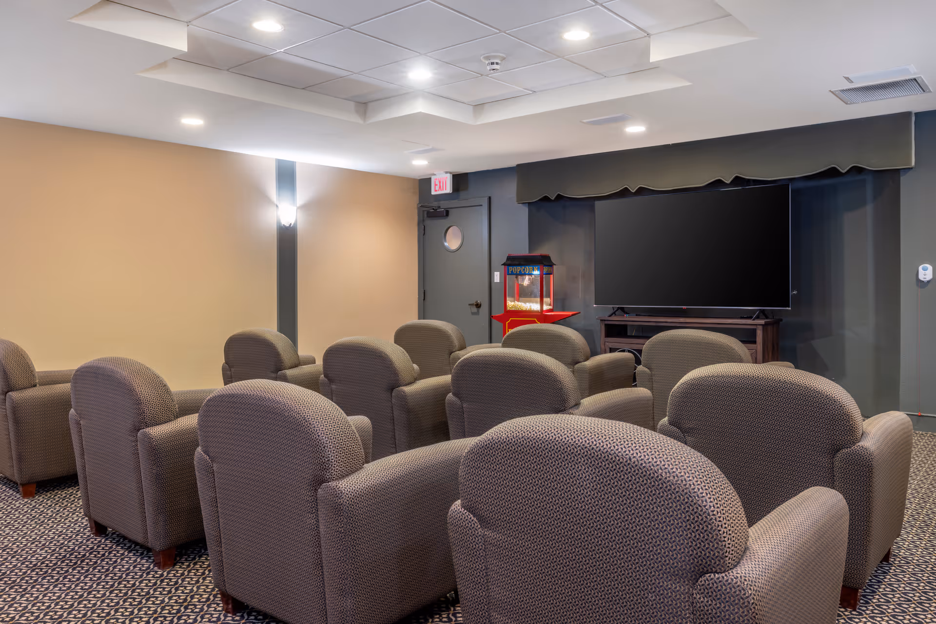 A small theater room with multiple rows of cushioned armchairs facing a large flat-screen TV mounted on a dark wall. A popcorn machine is placed next to the TV, and the room has beige walls, patterned carpet, and recessed ceiling lights.