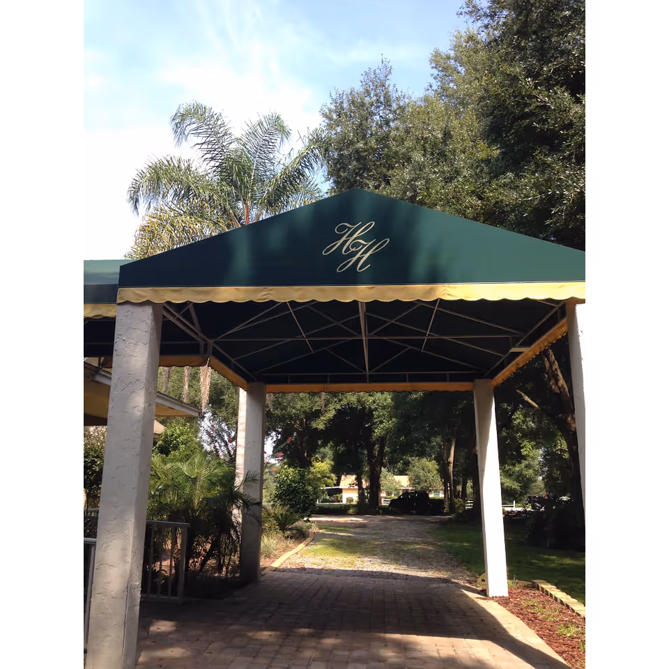 Green entrance canopy monogrammed 'HH' over a brick driveway leading into a tree-lined property.