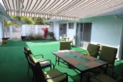 Covered outdoor patio with green cushioned seating, a dining table, striped retractable awning, and artificial turf.
