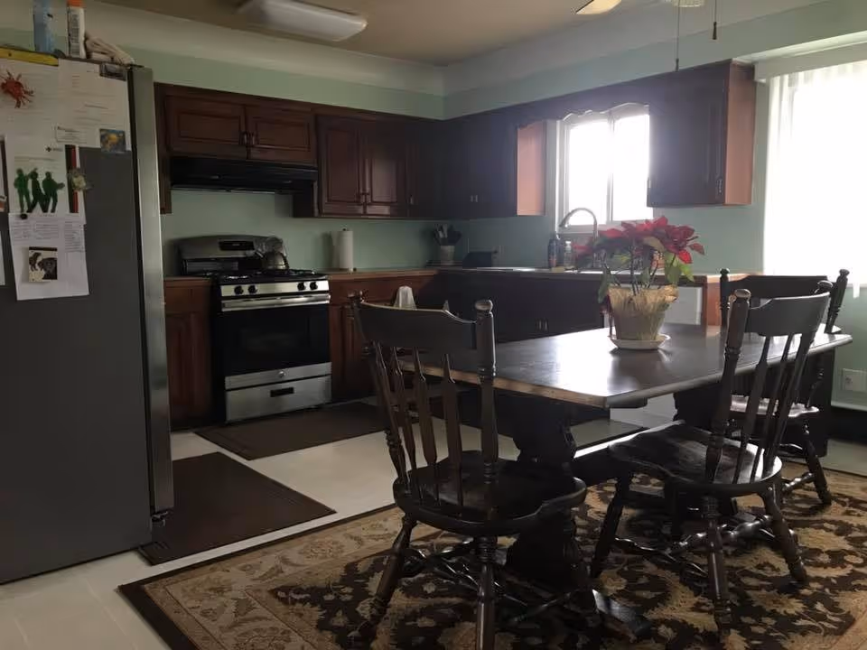 A kitchen and dining area with dark wooden cabinets, a stainless steel stove and refrigerator, a wooden dining table with four chairs, a floral centerpiece on the table, and a window letting in natural light.