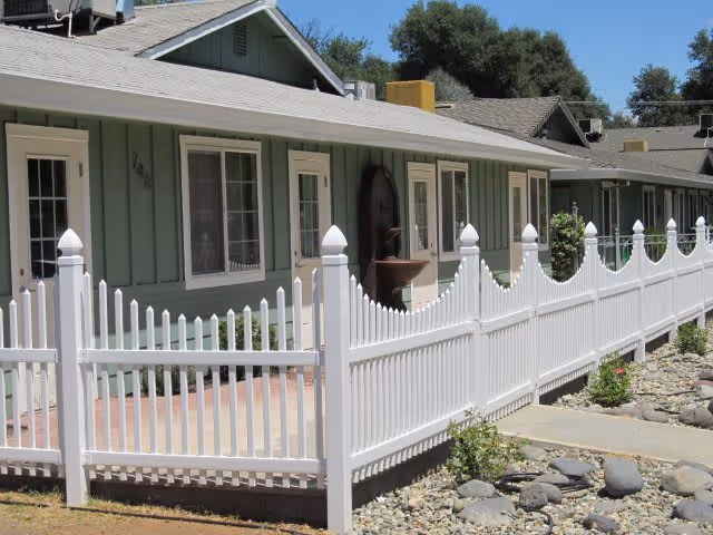 Exterior view of a single-story residential building with green siding and multiple windows, surrounded by a white picket fence. There is a rocky landscaped area with some small plants in front of the fence, and trees are visible in the background under a clear blue sky.