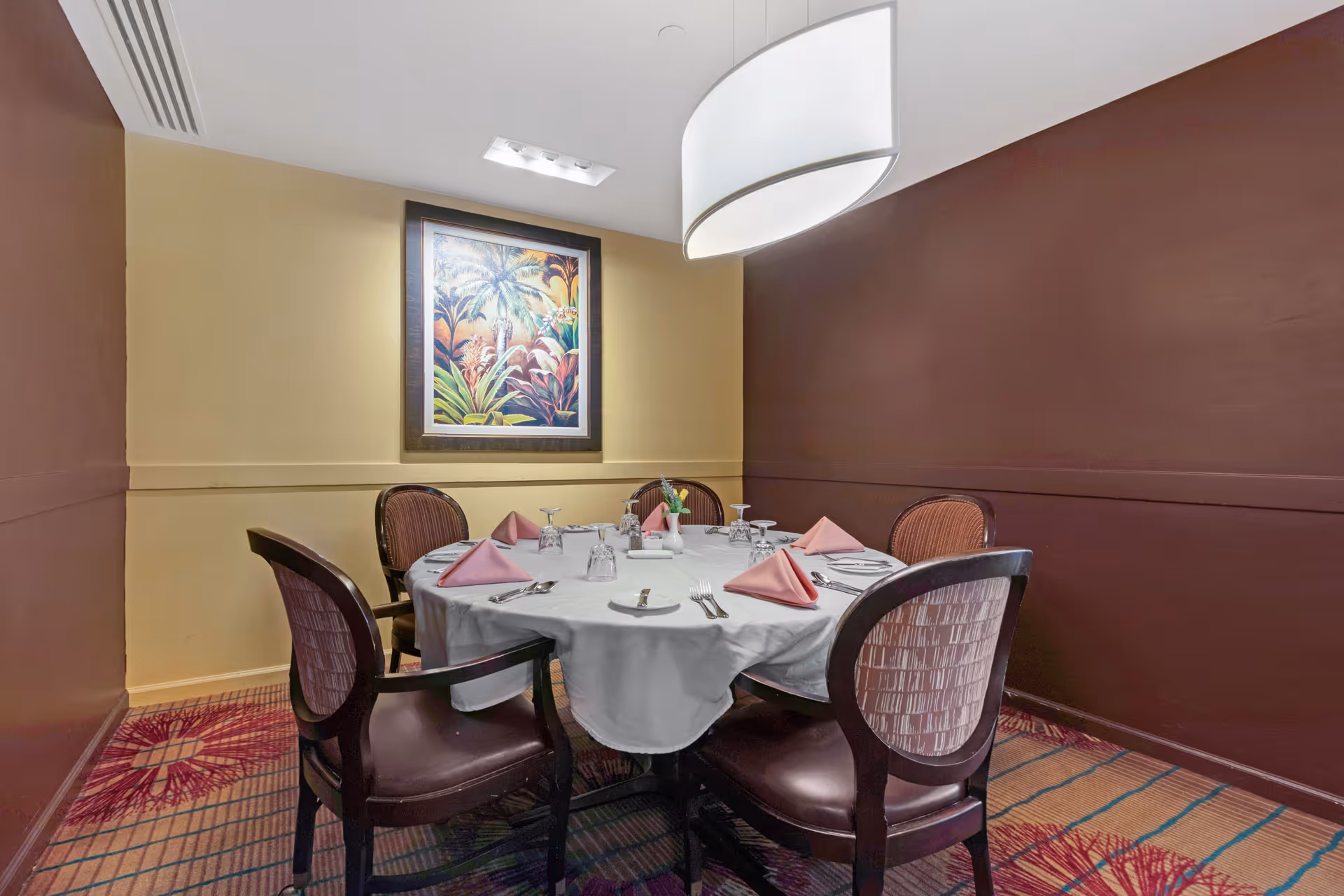 A small dining room with a round table covered with a white tablecloth, set with pink folded napkins, plates, glasses, and silverware. Four wooden chairs with cushioned seats surround the table. The walls are painted in two tones, beige and dark brown, and a framed tropical-themed painting hangs on the beige wall. A modern white pendant light hangs above the table, and the carpet has a colorful pattern.