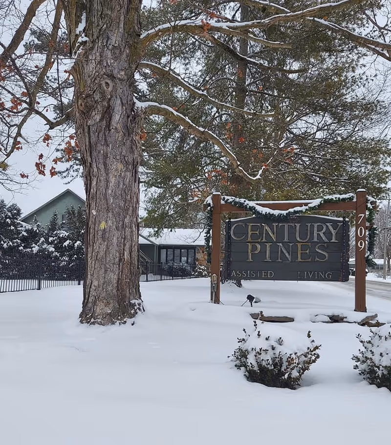 Snow-covered outdoor scene featuring a large tree and a wooden sign for Century Pines Assisted Living, with a building and fence in the background.