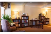 A cozy senior living common room with bookshelves, armchairs, a side table, lamps, and warm lighting.
